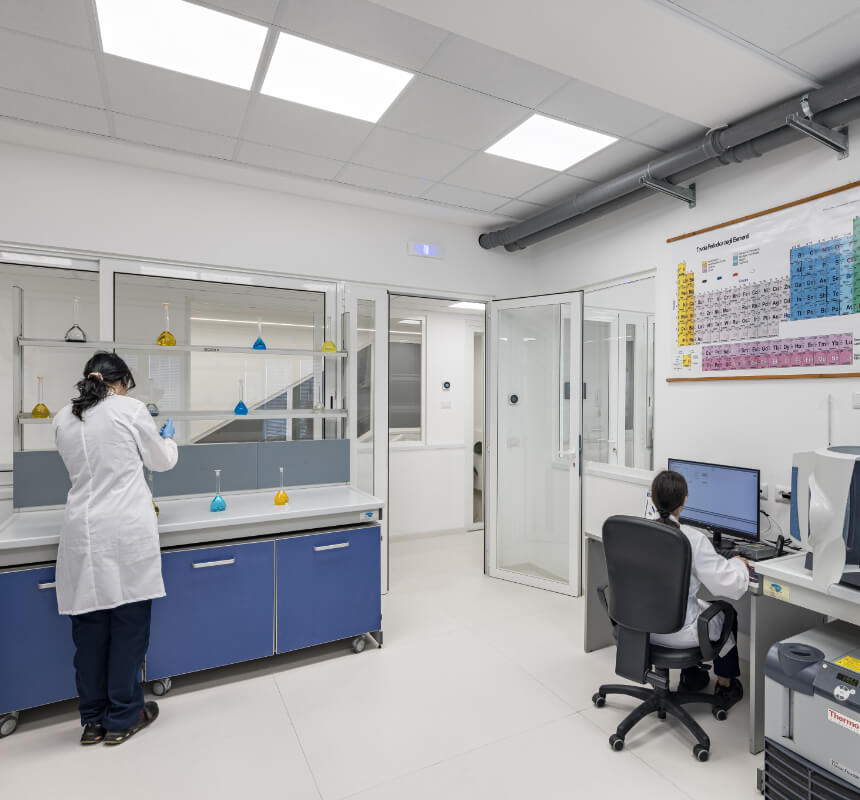 Two people in lab coats work in a modern laboratory; one stands at a counter with flasks, while the other sits at a computer. The room is bright, with lab equipment and a periodic table on the wall.