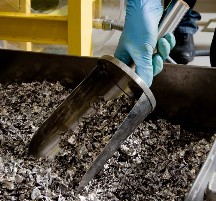 Una mano guantata regge una grande paletta metallica riempita di materiale triturato di aspetto metallico in un vassoio