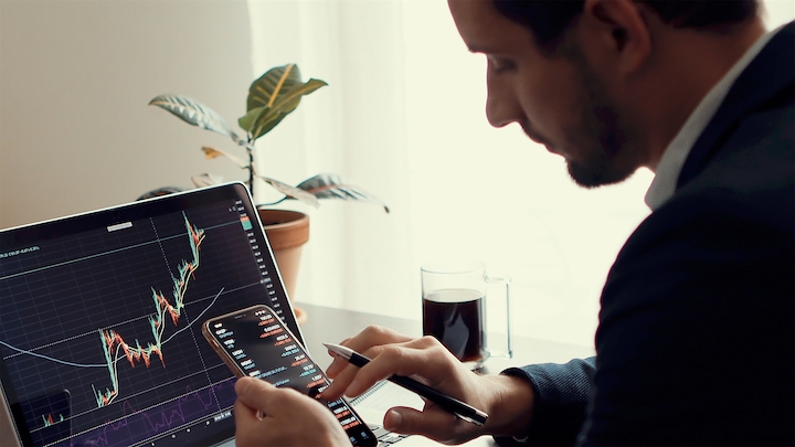 A man in business attire analyzes stock market charts on a laptop while holding a smartphone. A potted plant, a glass mug with a dark beverage, and a notepad are on the desk in a well-lit room.