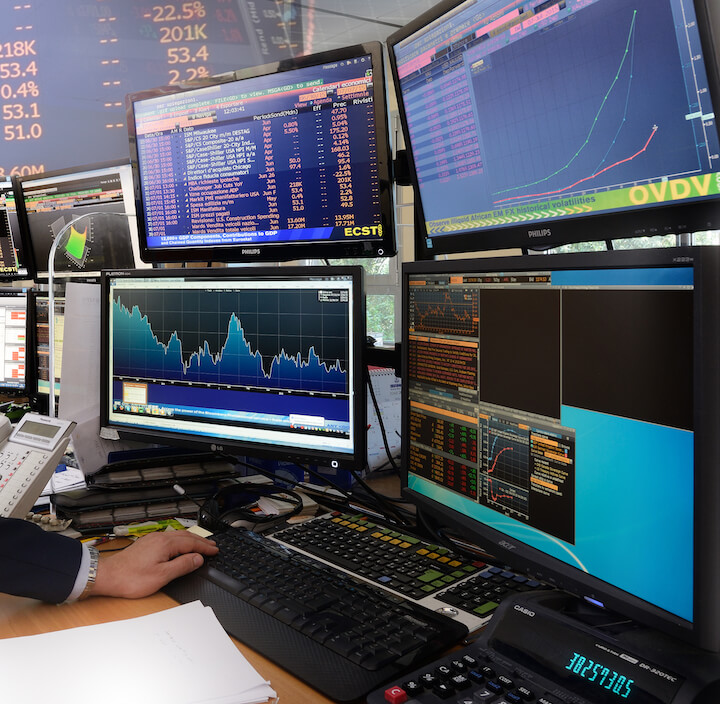 A person works at a desk with multiple computer monitors displaying stock market charts, financial data, and graphs in a modern trading office environment.