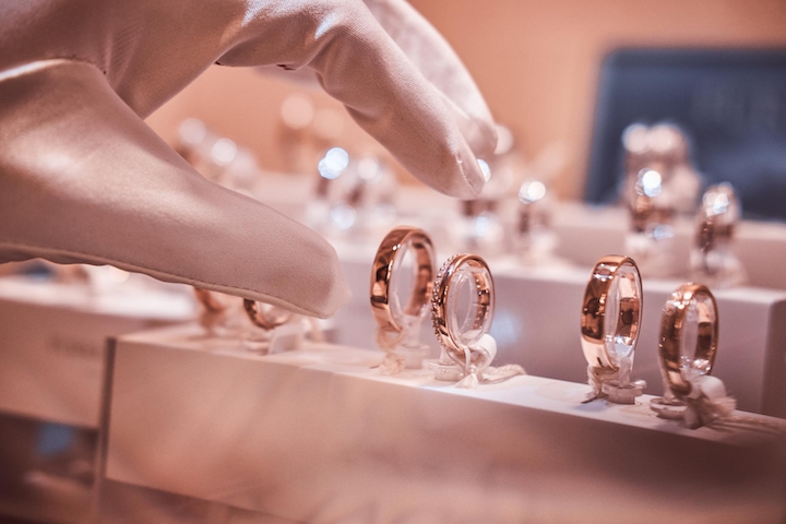 A gloved hand reaches for a gold ring among several rings displayed in a jewelry store showcase, with a soft, elegant lighting highlighting the jewelry.
