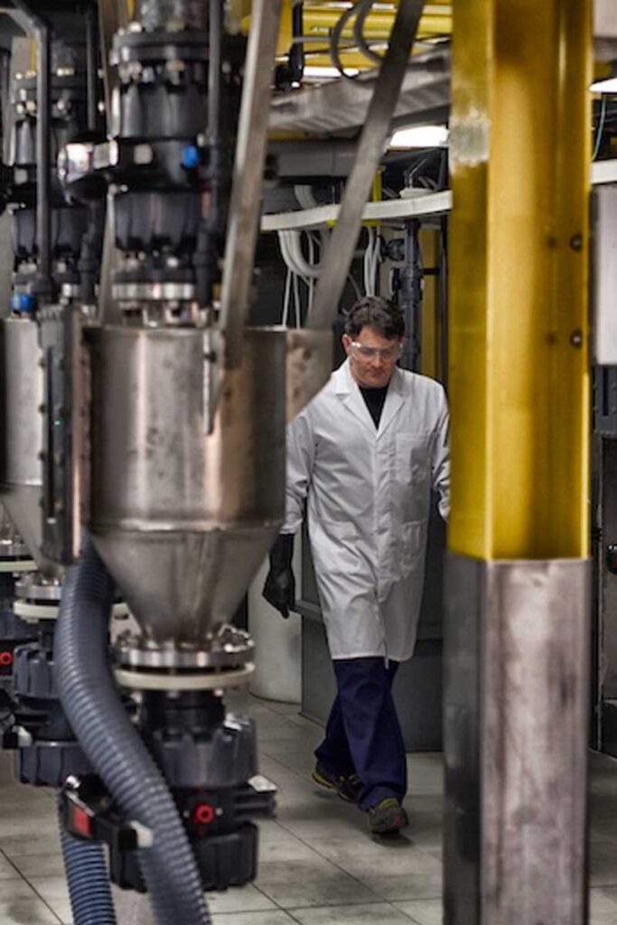 Una persona in camice bianco e occhiali di sicurezza attraversa una struttura industriale con grandi macchinari e tubi metallici.