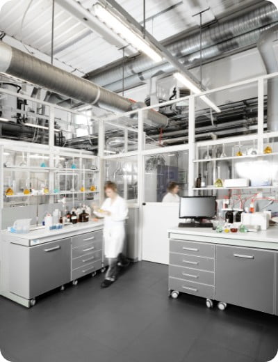 A bright, modern laboratory with stainless steel workstations, shelves of glassware, bottles, and equipment. Two people in white lab coats move around, creating a sense of activity and research.