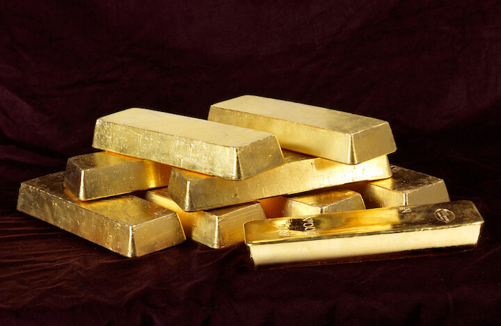 A stack of shiny gold bars is arranged on a dark, rich-colored surface, with several bars lying on top of each other and one bar slightly separated in the foreground.
