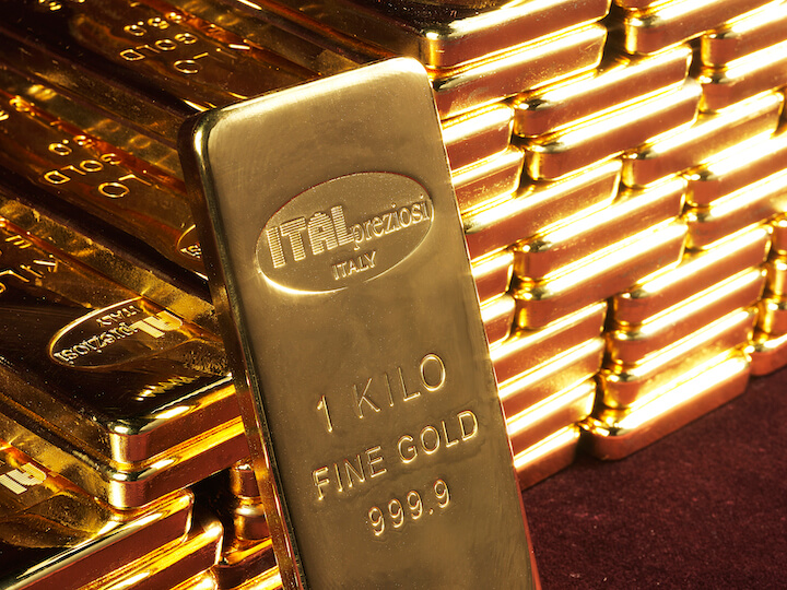 A close-up of a one-kilogram gold bar labeled ITALPREZIOSI ITALY 1 KILO FINE GOLD 999.9 stands upright in front of a large stack of similar shiny gold bars.