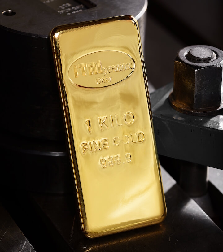 A shiny 1-kilogram gold bar stamped with ITALpreziosi ITALY, 1 KILO FINE GOLD, and 999.9 rests against industrial metal machinery in a dimly lit setting.