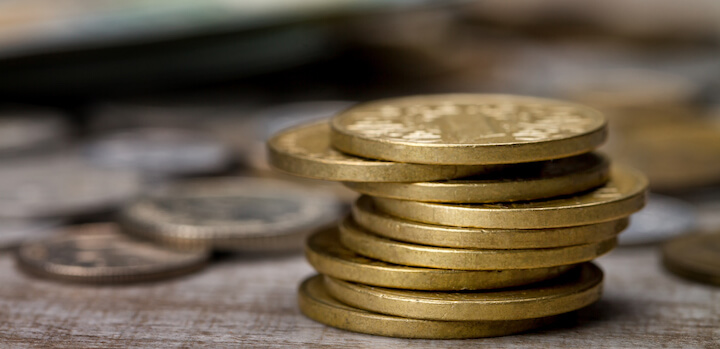 A close-up of several stacks of gold coins placed on a wooden surface, with more coins blurred in the background.
