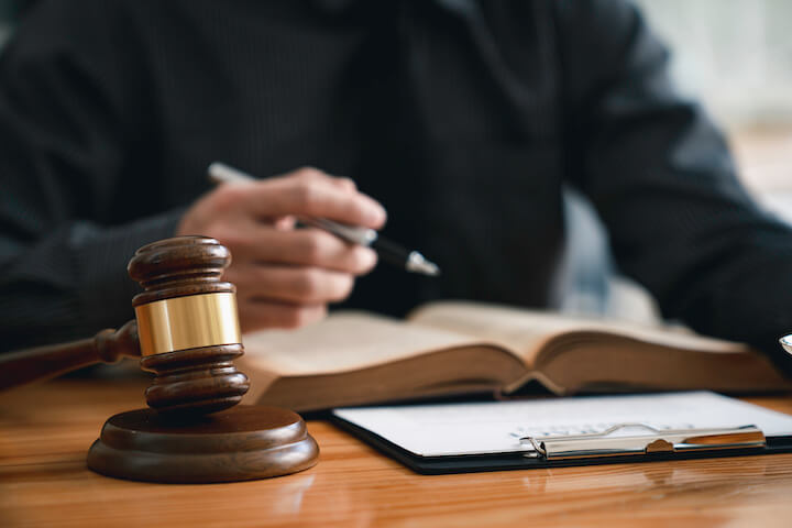 A person in dark clothing sits at a desk with an open book, holding a pen. A wooden judge’s gavel and a clipboard with papers are on the desk in the foreground.