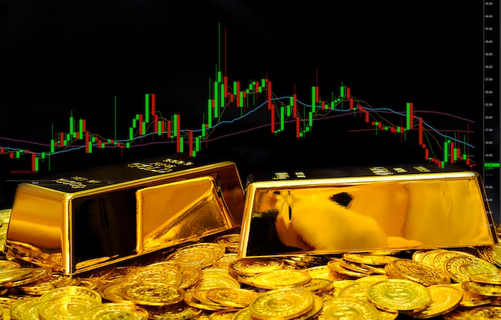 Two gold bars and several gold coins are displayed in the foreground, with a colorful stock market candlestick chart visible in the background on a black screen.