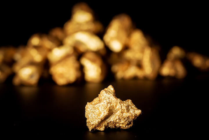 A single gold nugget is in sharp focus on a dark surface, with a blurred pile of additional gold nuggets in the background against a black backdrop.