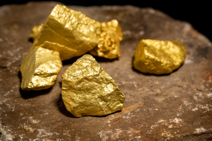 Four shiny gold nuggets rest on a rough brown stone surface, with their irregular shapes and metallic luster standing out against the earthy background.