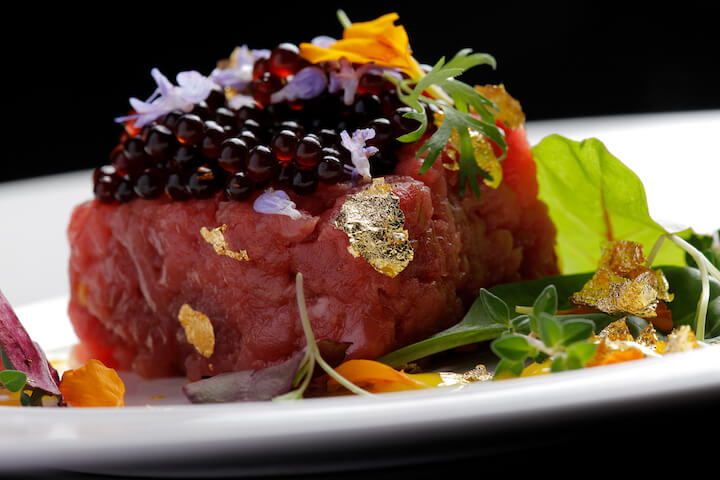 A close-up of gourmet steak tartare topped with caviar, edible flowers, microgreens, and gold leaf, artfully plated on a white dish against a black background.