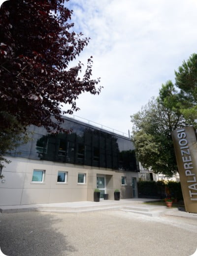 Modern office building with dark glass windows and light facade, surrounded by trees. A tall metallic sign reading ITALPREZIOSI stands at the entrance. The sky is partly cloudy.