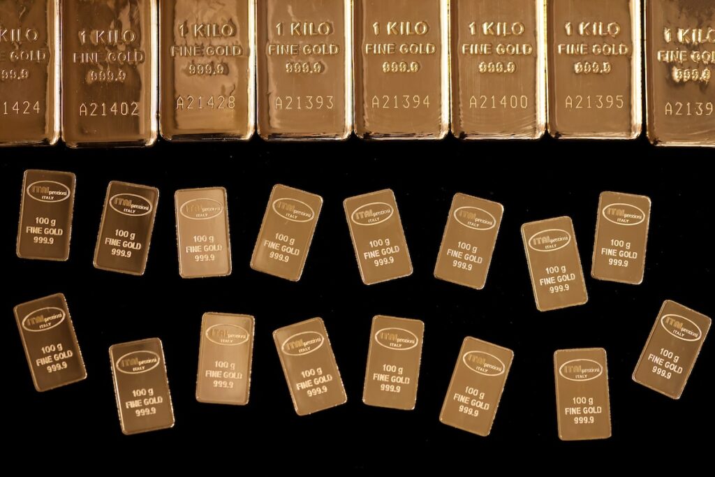 Rows of shiny gold bars are arranged on a black surface. The back row features large 1-kilo bars, while the front rows display smaller 100-gram bars, each stamped with weight and purity markings.